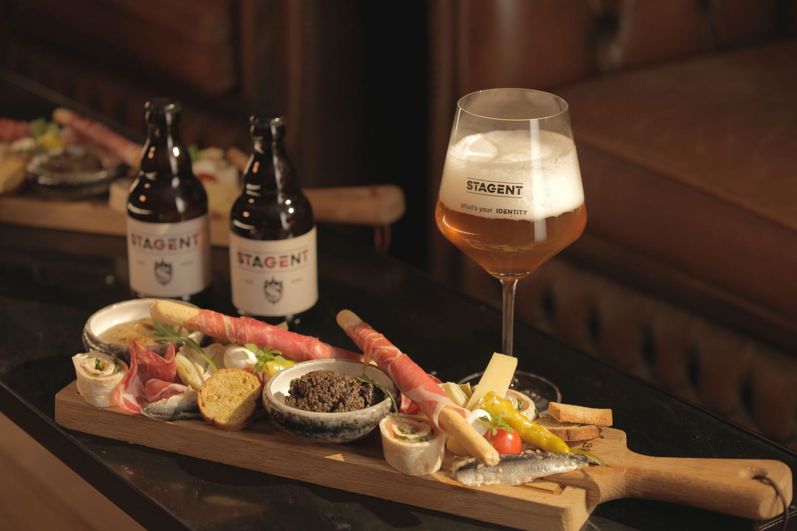 Een rijkgevulde borrelplank met charcuterie, kaas en hapjes, naast een gevuld Stagent-glas en twee flesjes.